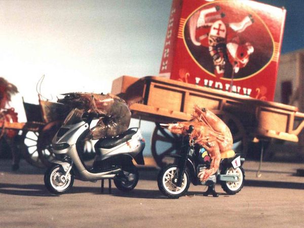 Twee garnalen zitten op speelgoedscooters voor een rood beschilderde kermiswagen.