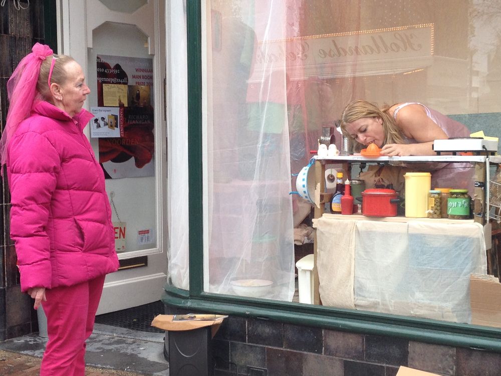 Een vrouw in een felroze jas kijkt door het glas naar de speelster die zich over haar keukentafeltje buigt.