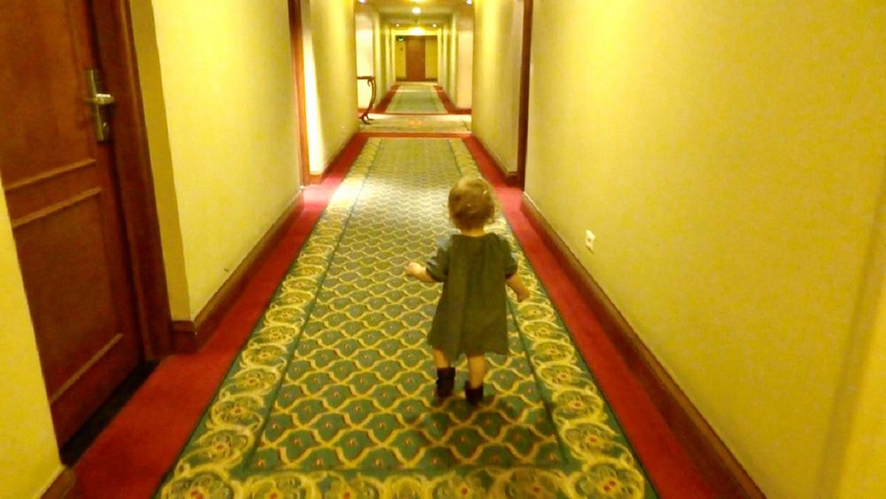 Videobeeld van een peuter in een groen jurkje die van de camera weg loopt door een lange hotelgang met gebloemd tapijt.