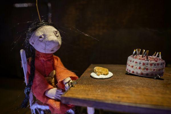 Close-up van een pop in rode kleren met warrig zwart haar aan een houten tafel; voor haar een koekje op een schoteltje en een taart met kaarsjes.