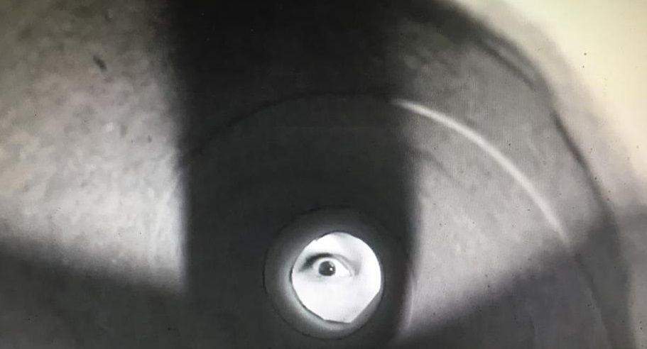 Filmbeeld door een deurspion: in het lichte rondje is een oog te zien dat terugkijkt.