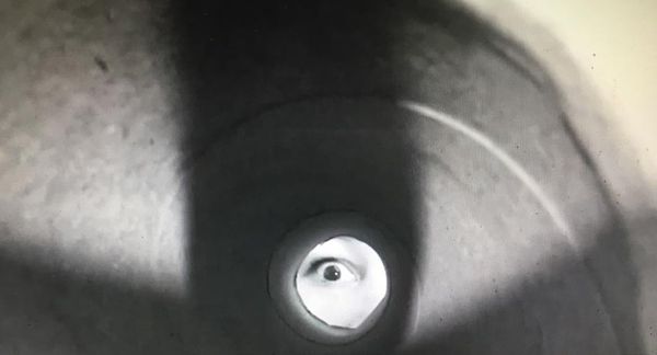 Filmbeeld door een deurspion: in het lichte rondje is een oog te zien dat terugkijkt.