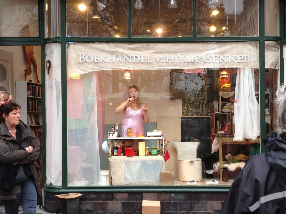Een vrouw in een roze jurk staat in de etalage van boekhandel Van Gennep achter haar keukentafeltje, twee voorbijgangers kijken toe.