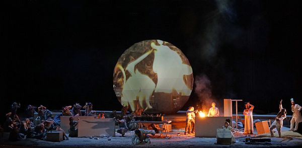 Toneelbeeld met de bol waarop dieren en figuren oplichten; op het podium zitten koorleden rond een vuurtje in de rook.