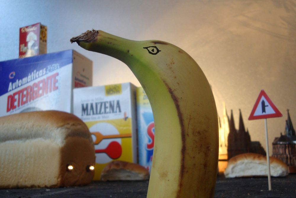 Een banaan met een getekend oog staat rechtop tussen pakken wasmiddel en maizena, met broden en een verkeersbord als stadsdecor eromheen.