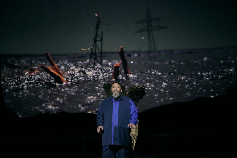 Michael König als Siegmund staat in het blauw voor de projectie van een ondergelopen landschap met hoogspanningsmasten.