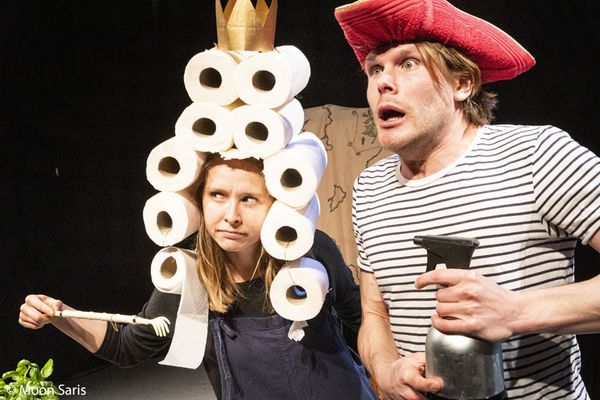 Twee spelers kijken verschrikt opzij: zij draagt een papieren kroon met een stapel wc-rollen op haar hoofd, hij een rode steek en een steelpan.