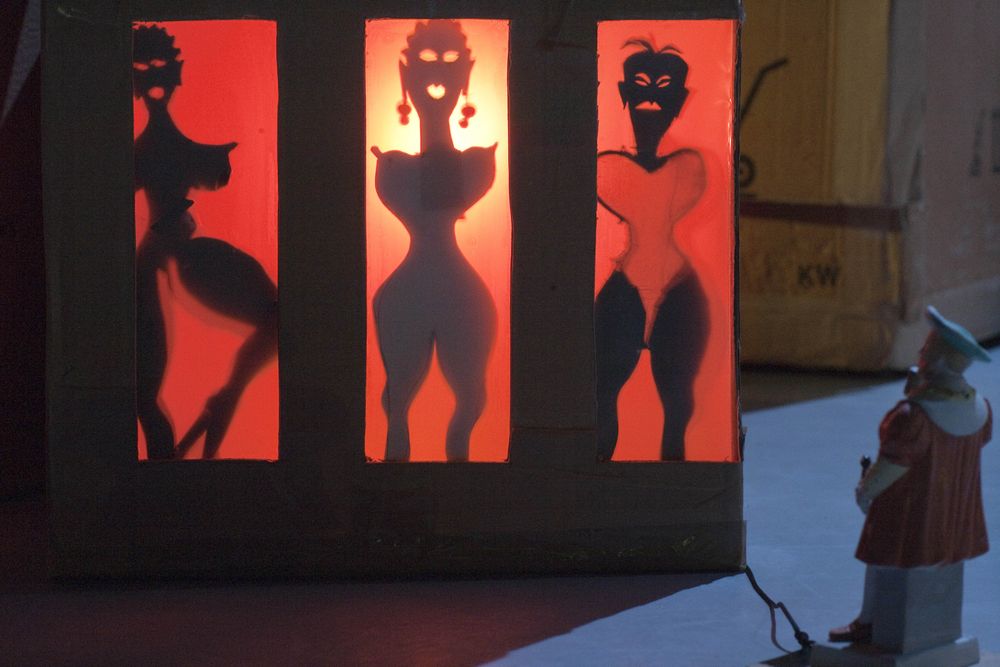 Een kartonnen kast met drie rood verlichte ramen waarin de silhouetten van vrouwen staan; ervoor kijkt een klein porseleinen poppetje toe.