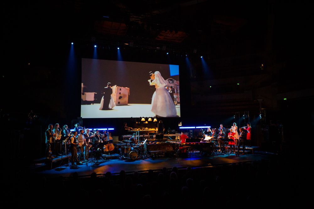 Concertzaal: op het scherm een bruidsfiguurtje in een lange sluier naast een kartonnen gebouw, daaronder speelt het ensemble.