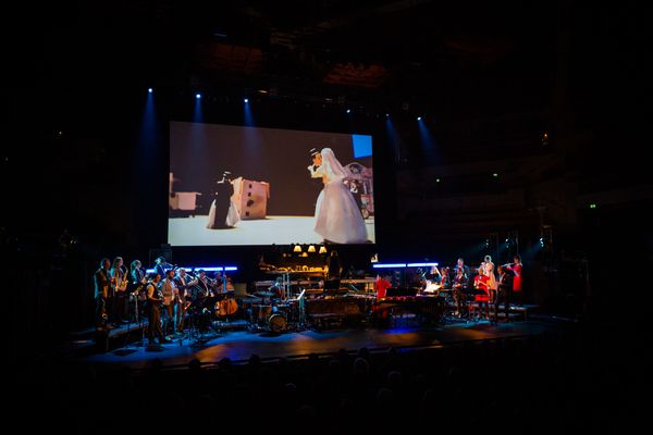 Concertzaal: op het scherm een bruidsfiguurtje in een lange sluier naast een kartonnen gebouw, daaronder speelt het ensemble.