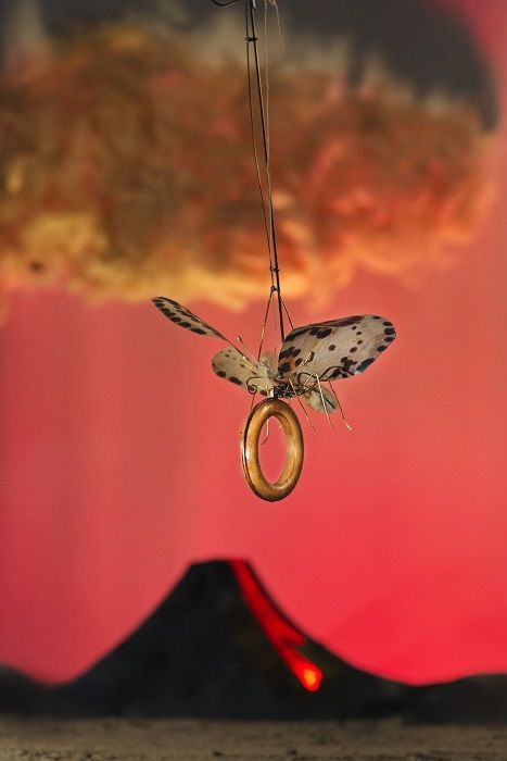 Een nachtvlinder hangt aan een draad met een gouden ring om zijn lijf; achter hem een rode lucht en een vulkaan met een gloeiende stroom lava.