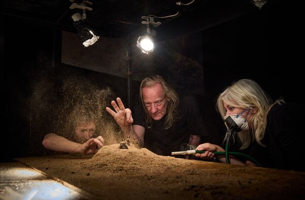 Drie makers gebogen over de zandmaquette in het licht van de werklampen; het opstuivende zand wordt door de spots gevangen.