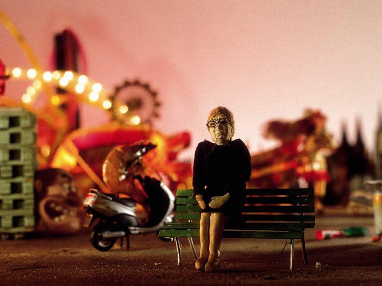 Een kleipoppetje van een oude vrouw zit 's avonds op een bankje op de kermis, met een scooter en verlichte attracties achter zich.