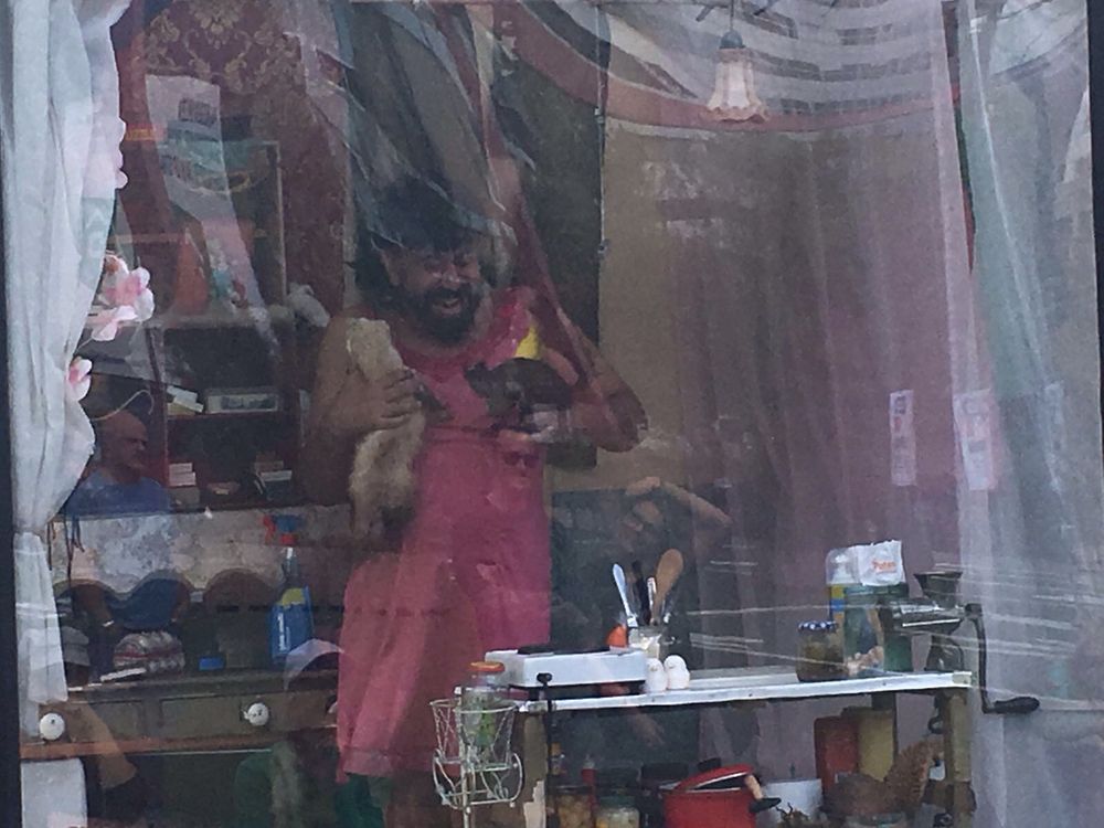 Een lachende speler in een roze jurk staat met een vogel in de handen in de etalage tussen keukengerei en potten.