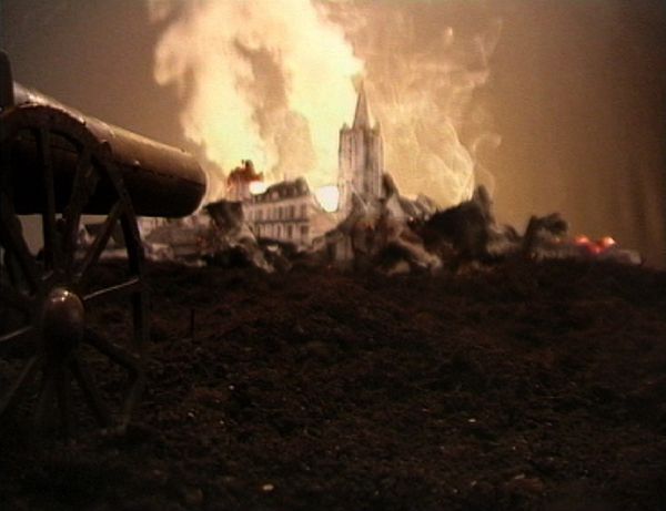 Een miniatuurstad staat in brand achter een aarden wal; de kerktoren steekt uit de vlammen, links op de voorgrond het wiel van een kanon.