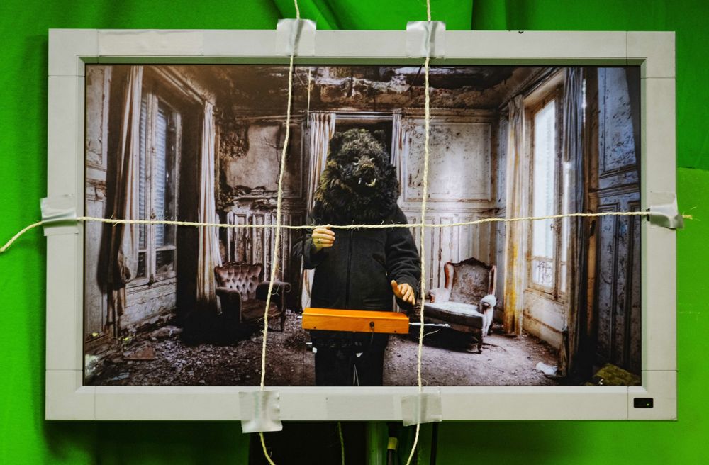 Een ingelijste foto van een vervallen kamer hangt met touwtjes voor een groen doek; op de foto staat een gestalte met een harig masker en een oranje kist.